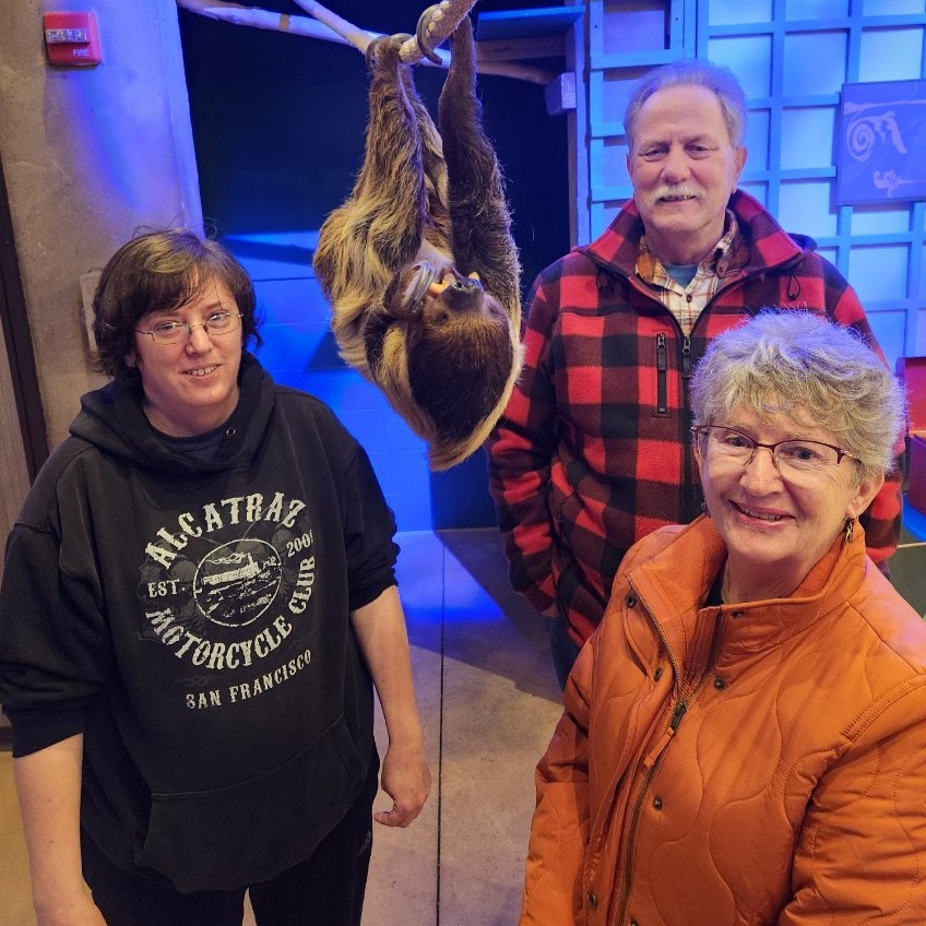 three people posing next to a sloth hanging from a branch