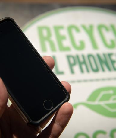 Photo showing a hand holding a phone, with a sign behind that reads "Recycle old phones here"