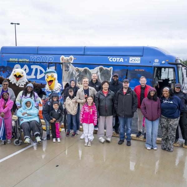 Group of 20 guests of all ages stand in front of the Zoo Bus smiling at the camera with the Zoo's Patriot Eagle and Sam Pelican Character Ambassadors
